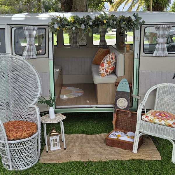 Darla the VW bus set up on the lawn with wicker chairs, a woven rug, and a picnic basket
